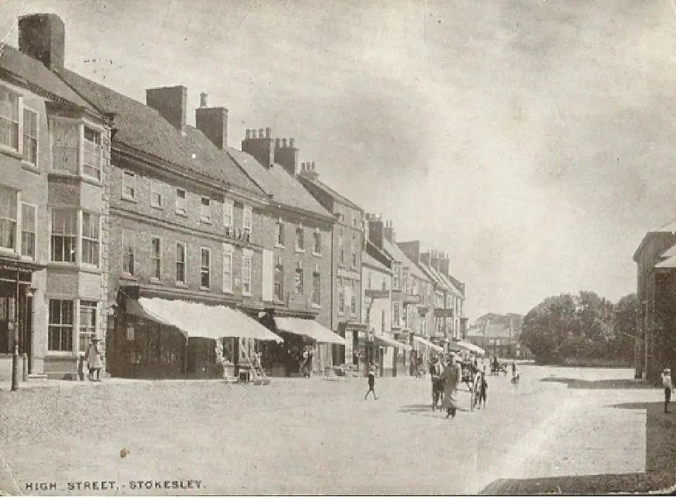Old postcard of Stokesley High Street: a long row of shops with awnings and a few people on a wide street.