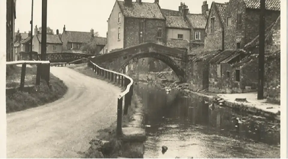 Old photograph of a stone arched bridge over the river beside a curving lane and houses.